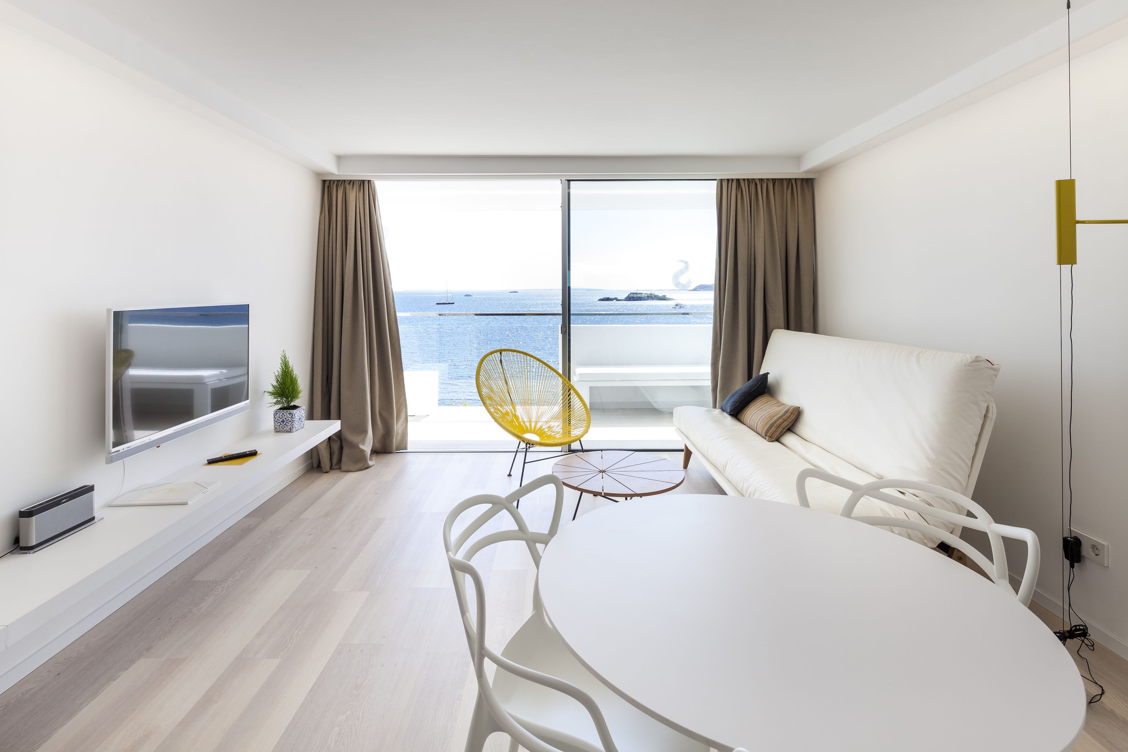 A modern living room with a large window, a flat-screen TV, a white sofa, a dining table, and a yellow chair.
