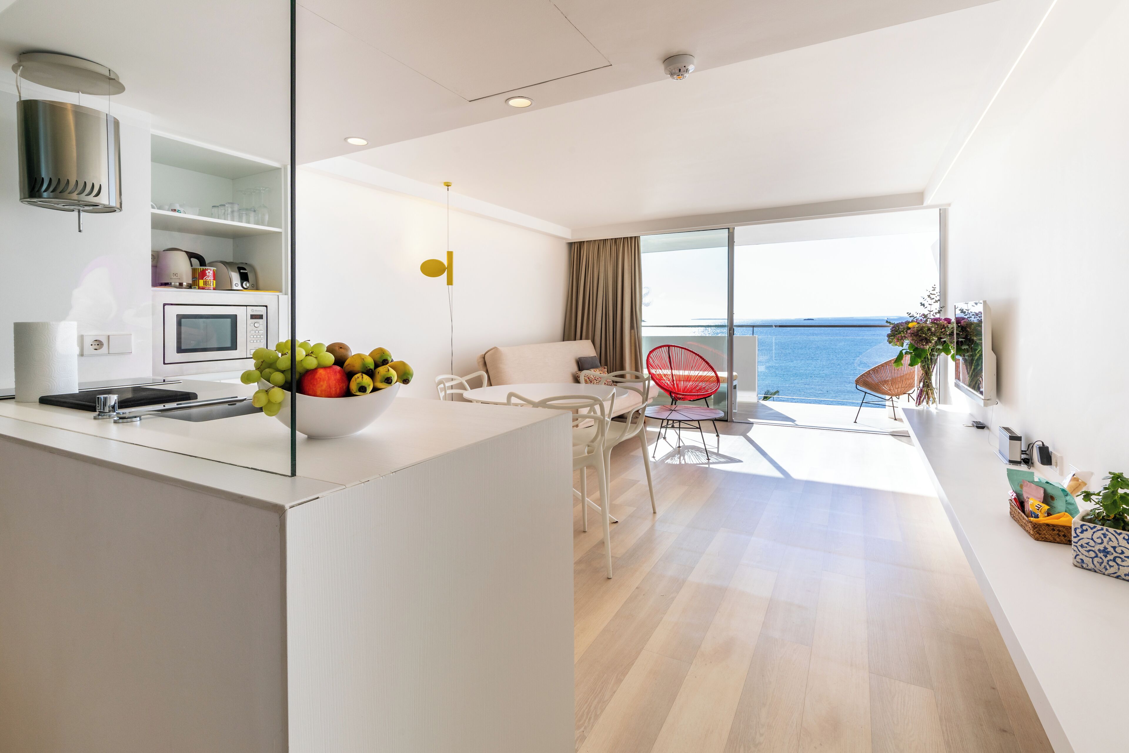 Una cucina moderna con forno a microonde, tostapane e una ciotola di frutta. C'è una zona pranzo con una sedia rossa e vista sul mare.