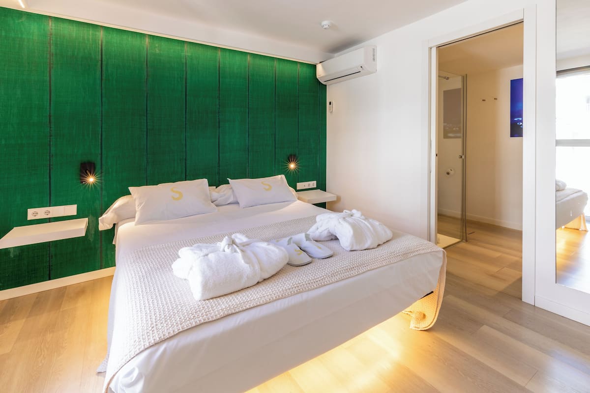 A modern hotel room with a large bed, green headboard, white bedding, and a glass shower area.