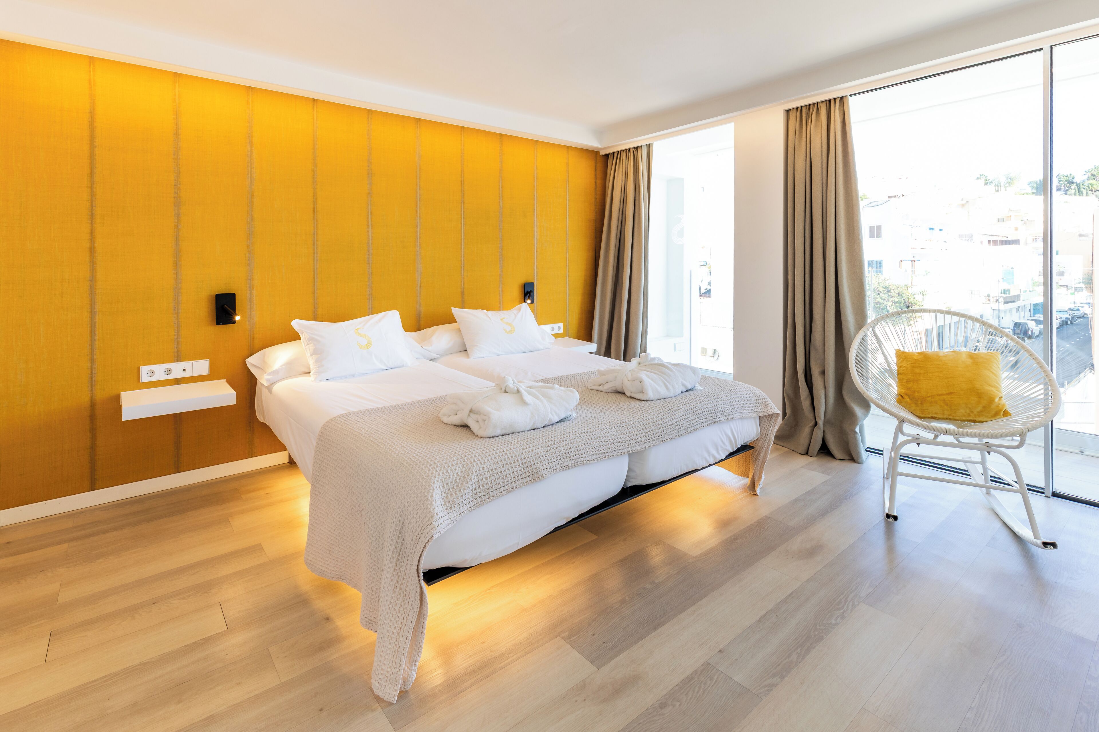 Habitación de hotel moderna con una cama grande, una pared de color amarillo brillante y vistas a la ciudad a través de amplias ventanas.