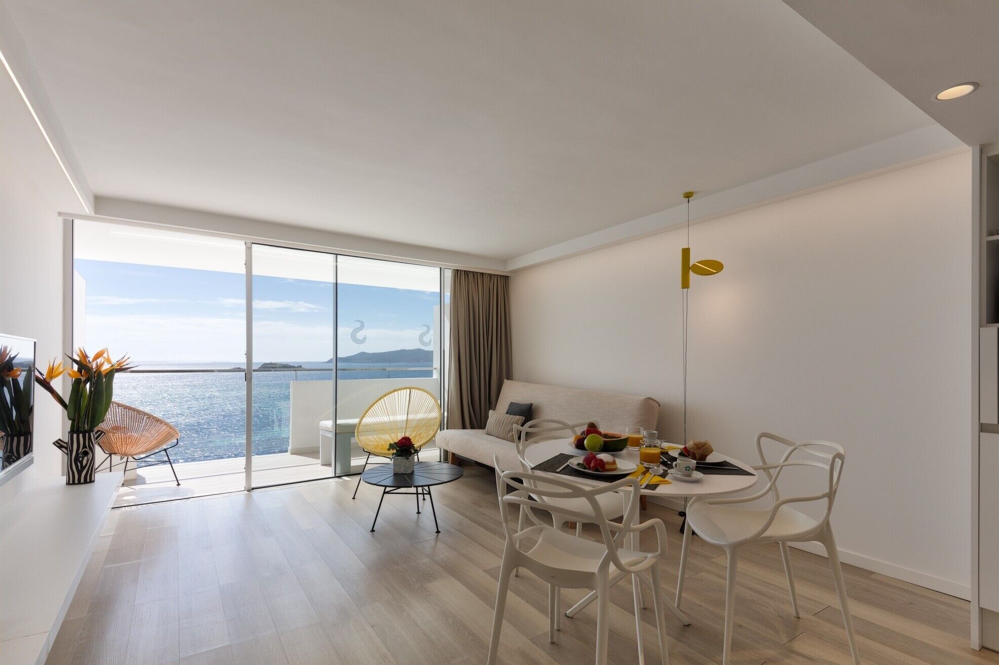 Una sala de estar moderna con un balcón con vistas al mar, un comedor con muebles blancos y una decoración minimalista.