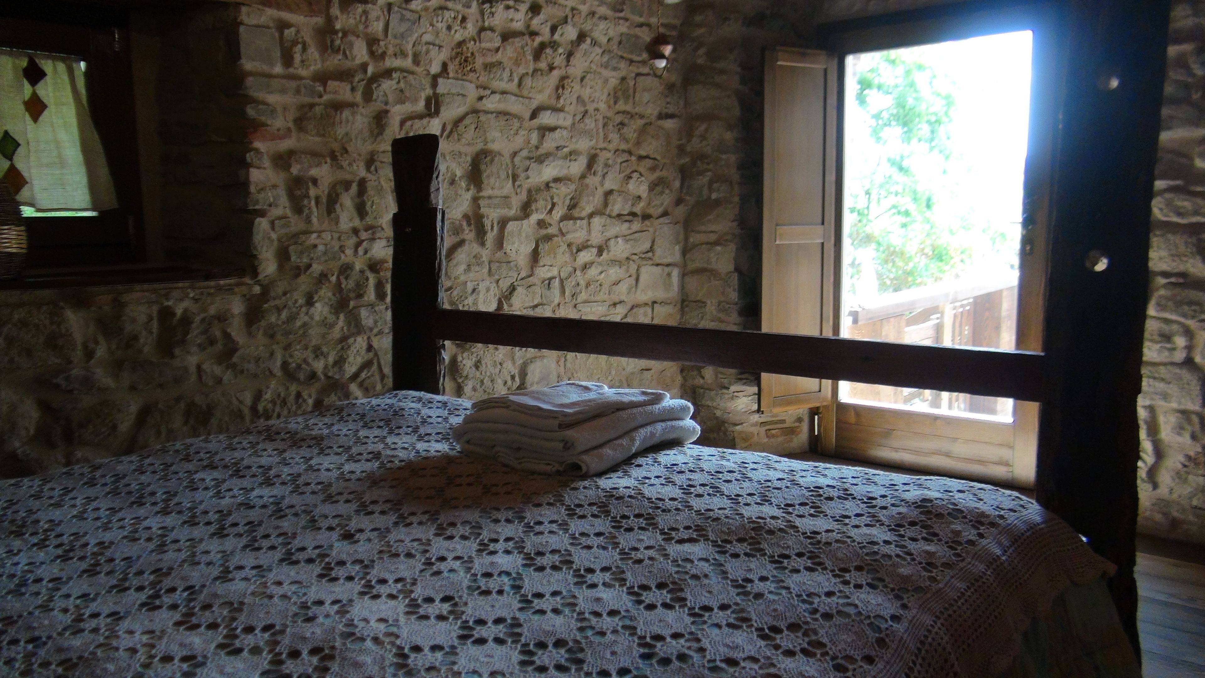 A room with a stone wall, a wooden bed frame, and a window with a view of greenery outside.