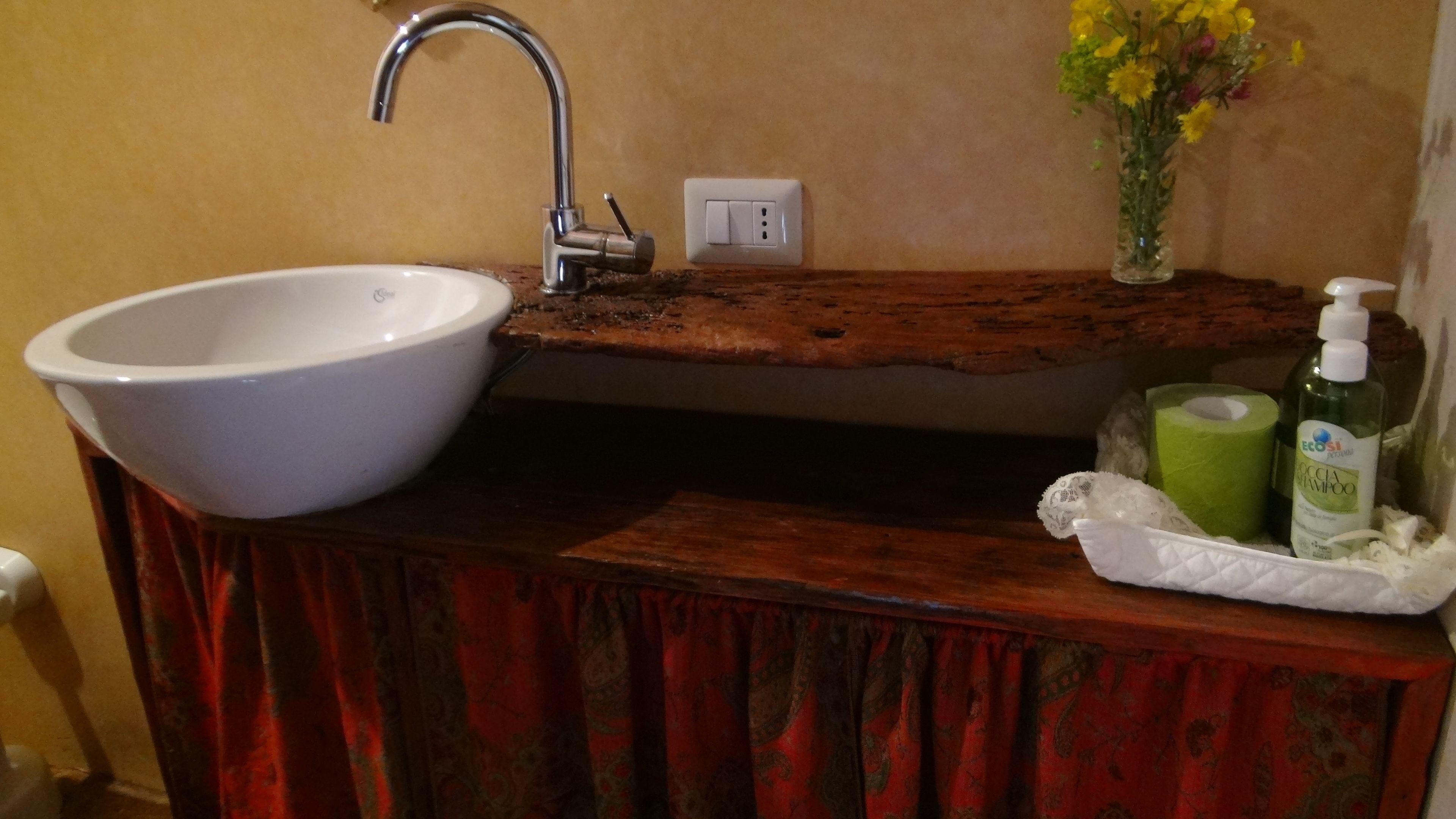 Une salle de bain avec un lavabo blanc, une étagère en bois, un vase avec des fleurs jaunes et des articles de toilette.