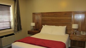 A hotel room with a bed, two bedside tables with lamps, a window with blinds, and a wooden headboard. - Reynolds Hotel (Lillooet)