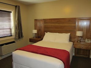 A hotel room with a bed, two bedside tables with lamps, a window with blinds, and a wooden headboard. - Reynolds Hotel (Lillooet)