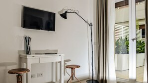 A modern living room with a flat-screen TV mounted on the wall, a white console table, two wooden stools, a floor lamp, and a sliding glass door leading to a balcony with plants. - Click&Sleep (Naples)