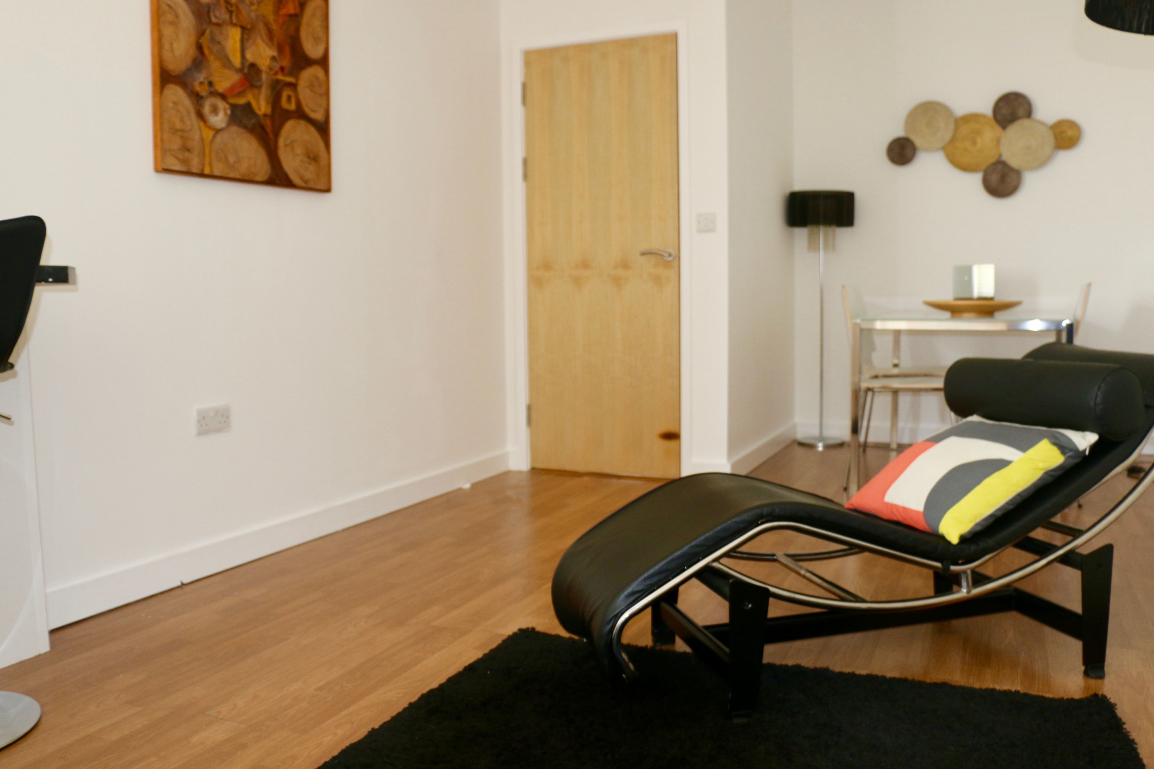 A modern living room with a black leather lounge chair, a wooden door, a small table with a lamp, and a wall art piece.