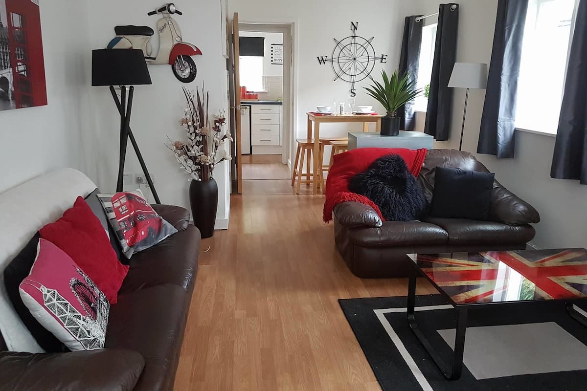A living room with a brown leather sofa, a red and white patterned table, and a wooden floor. There is a dining area with a table and chairs in the background.