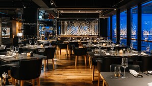 A modern restaurant with tables set for dining, a bar area, and large windows offering a view of the cityscape. - MACq 01 Hotel (Hobart)