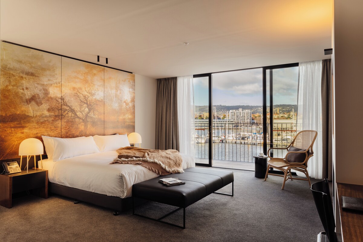 A modern hotel room with a large bed, a view of the city, a chair, and a desk.