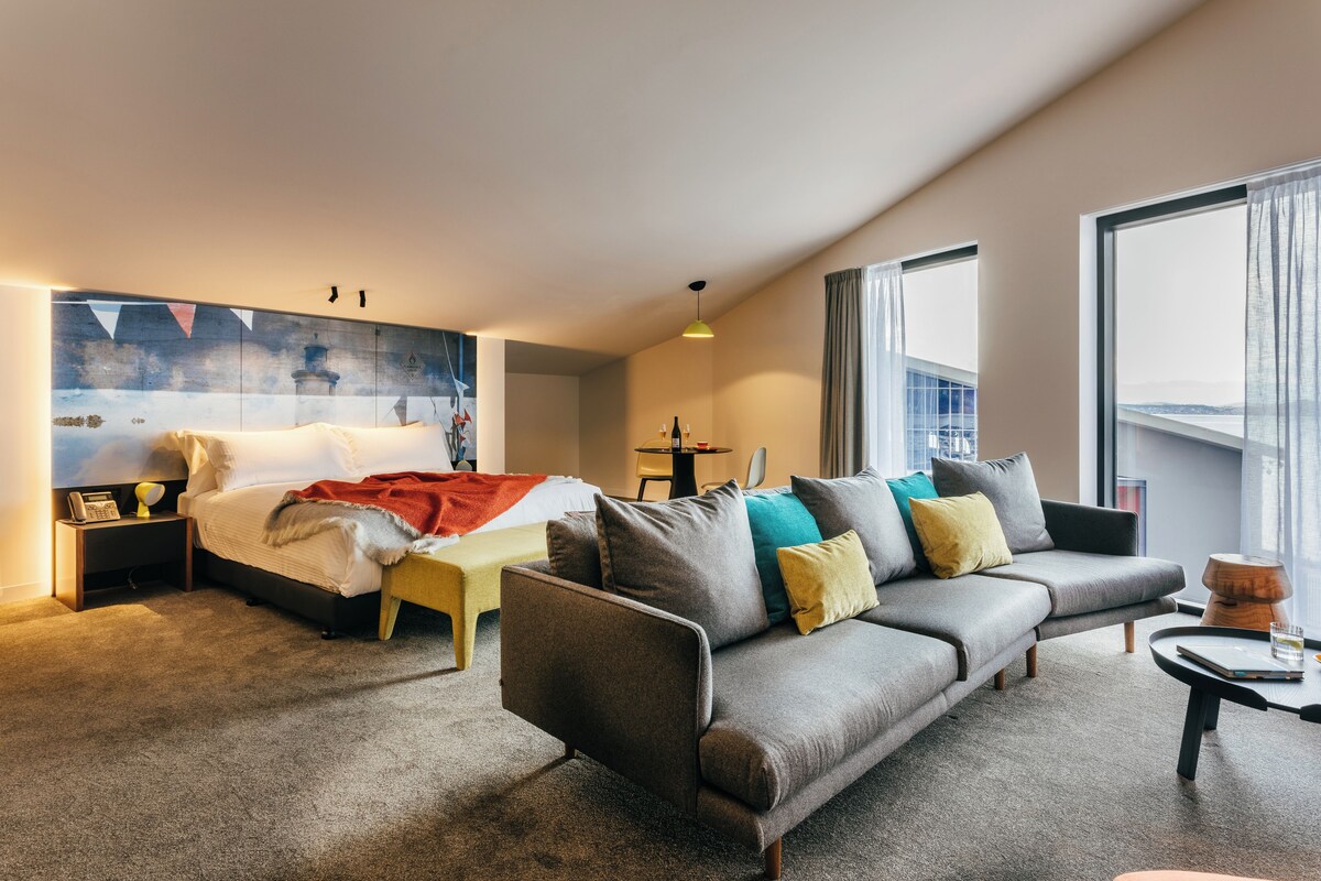 A modern hotel room with a bed, sofa, and a large window.