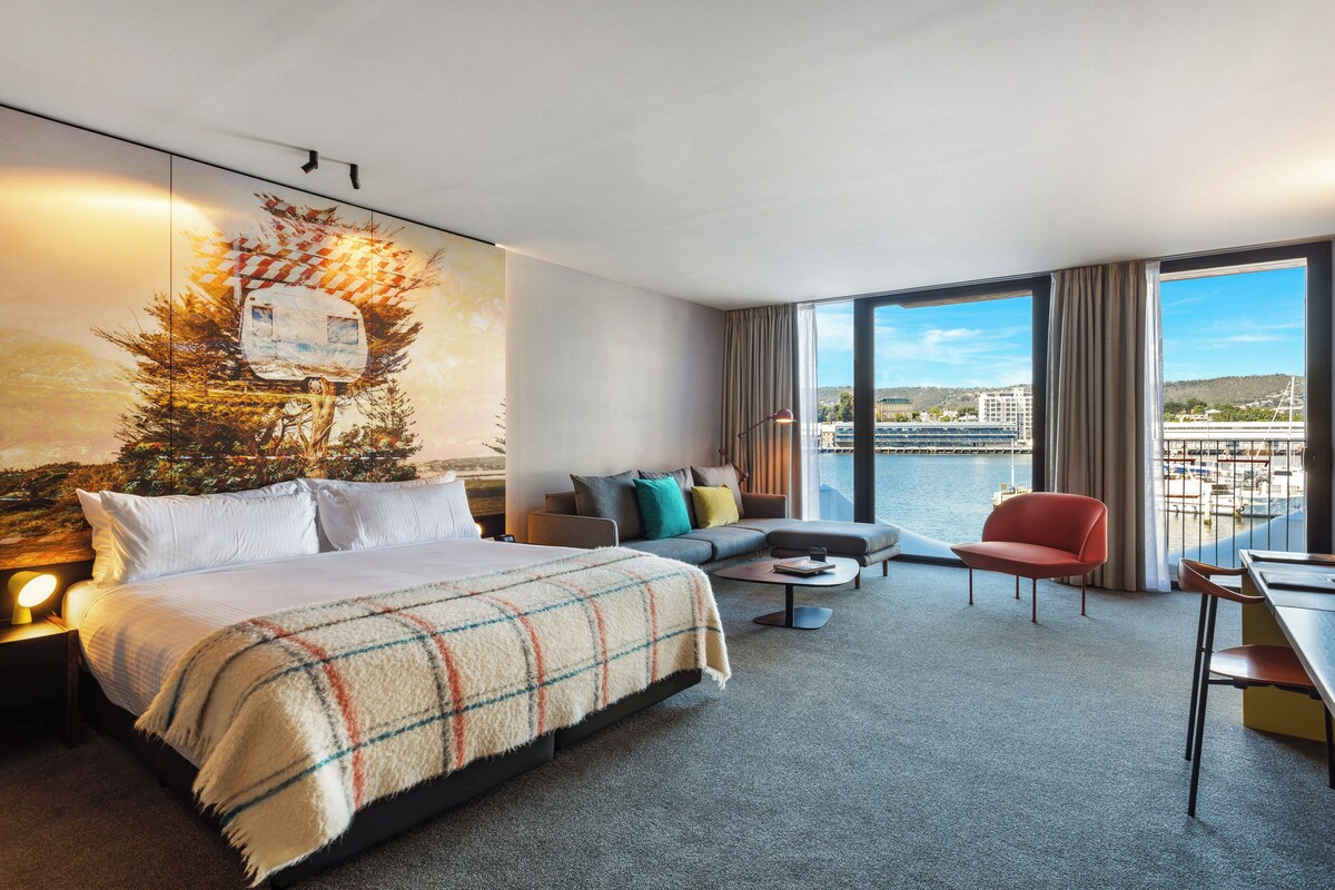 A hotel room with a bed, a sofa, a dining table, and a view of the city and water.