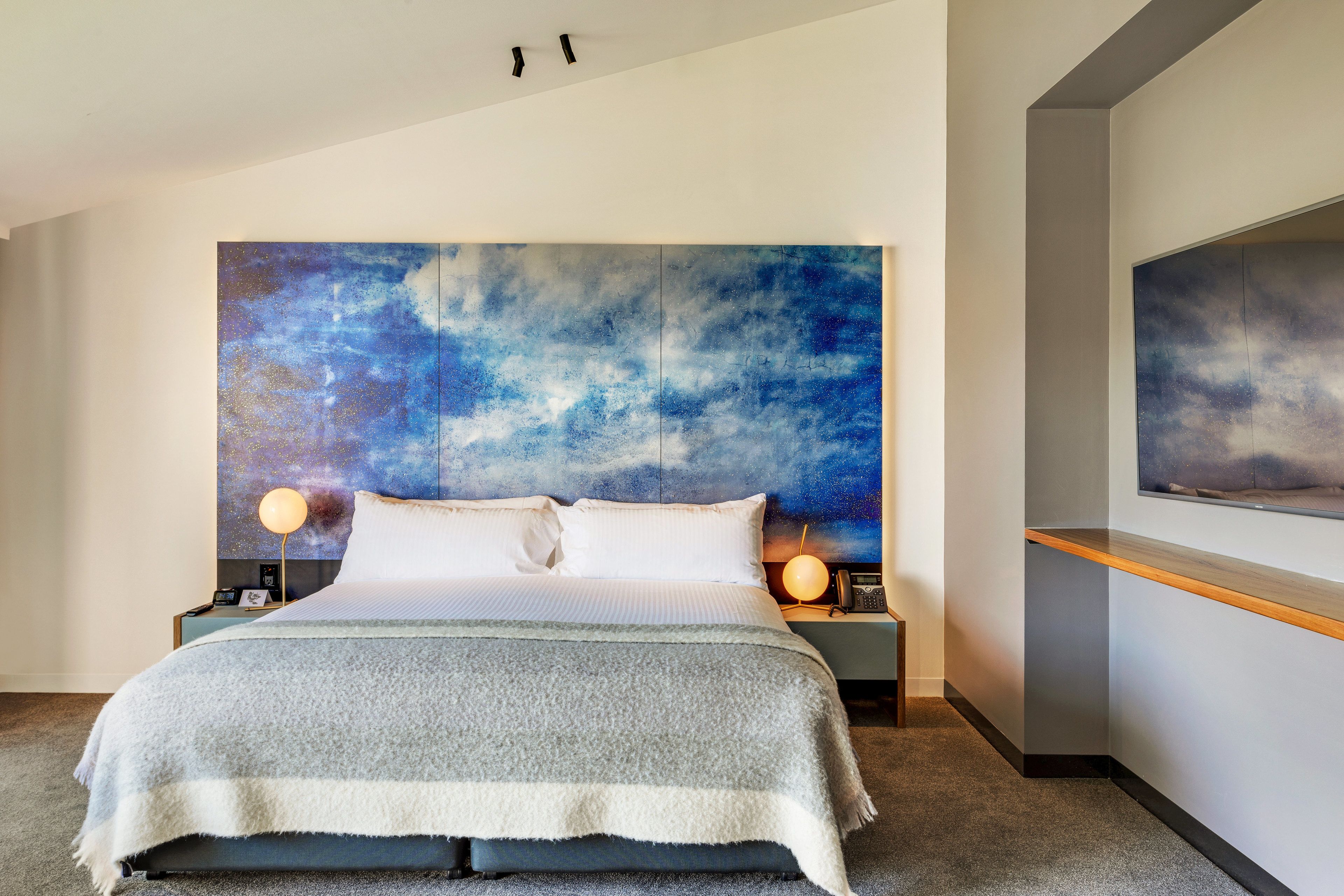 A modern bedroom with a large bed, two bedside tables, a wall-mounted TV, and a large abstract painting above the bed.