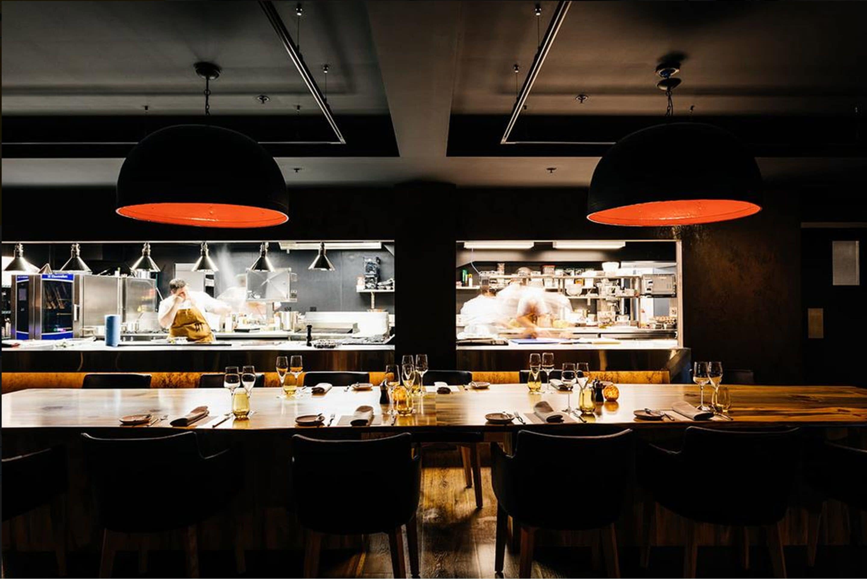 Un restaurante con una barra larga, varias mesas e iluminación colgante.