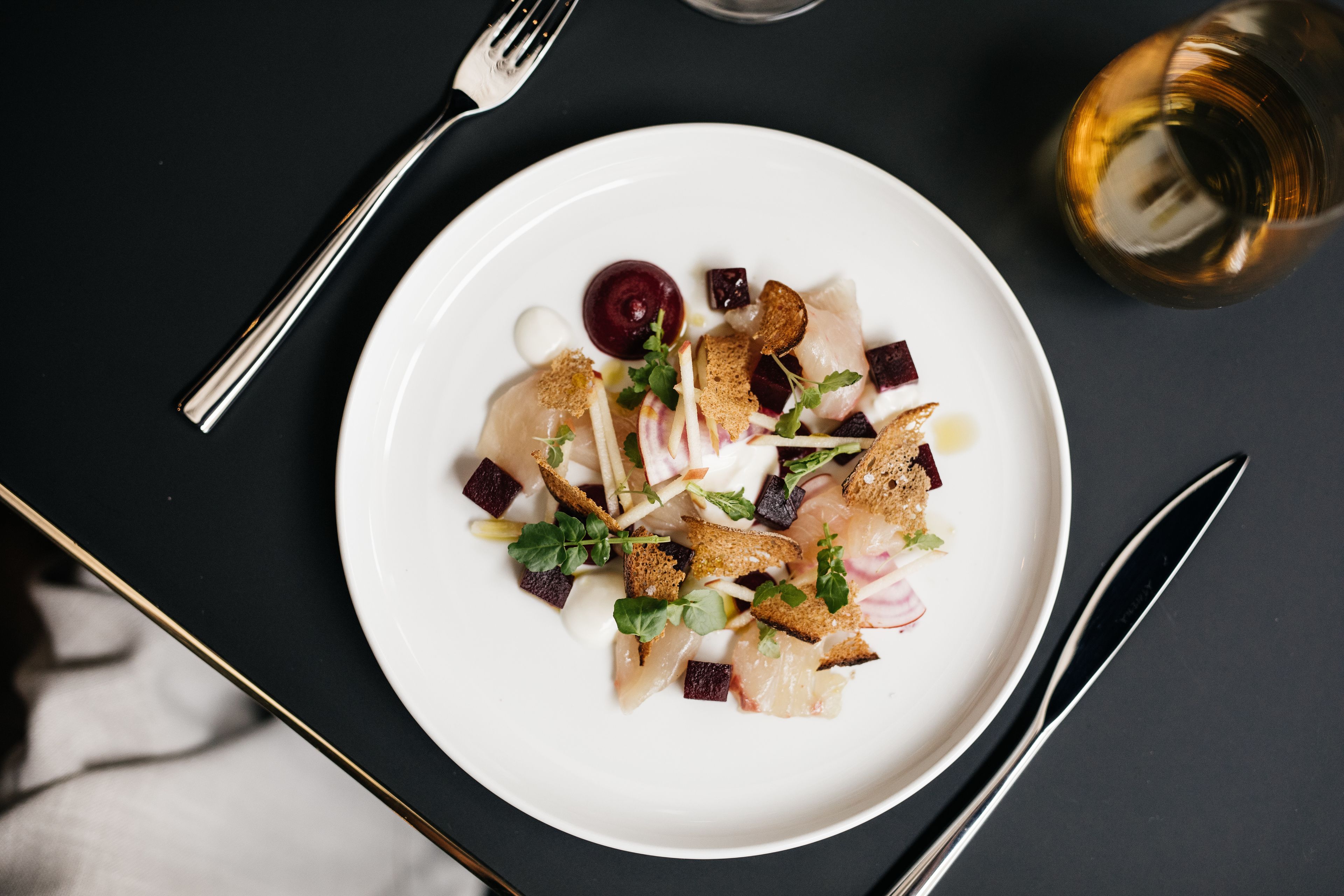 A white plate with a salad, accompanied by a fork and knife on a dark surface.