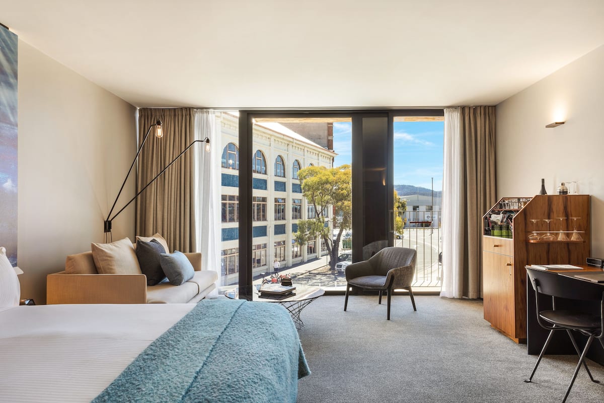 Quarto de hotel com uma cama grande, uma escrivaninha, uma cadeira, um sofá e vista para a cidade através de uma porta de vidro.