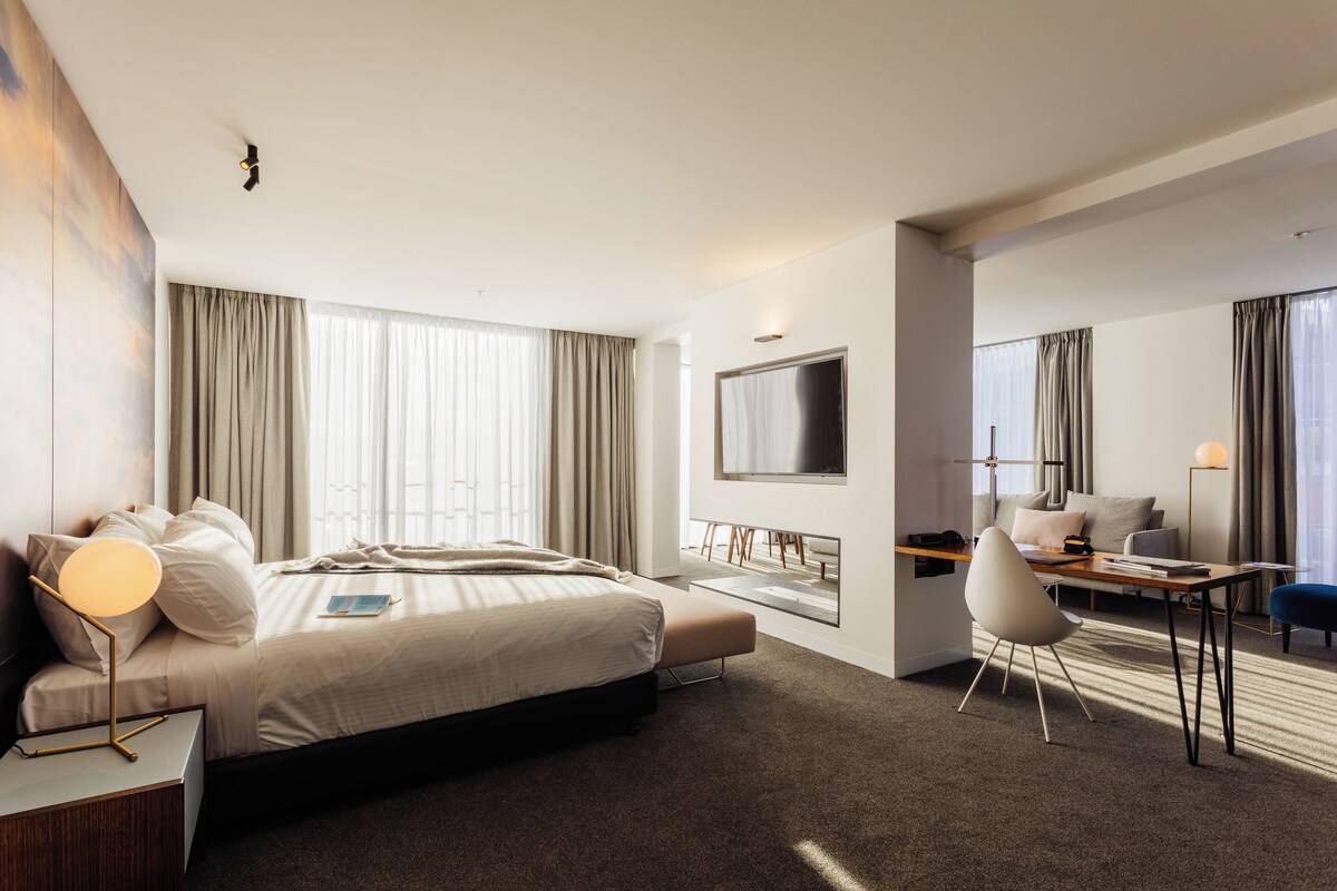A modern hotel room with a bed, desk, chair, TV, and large windows.