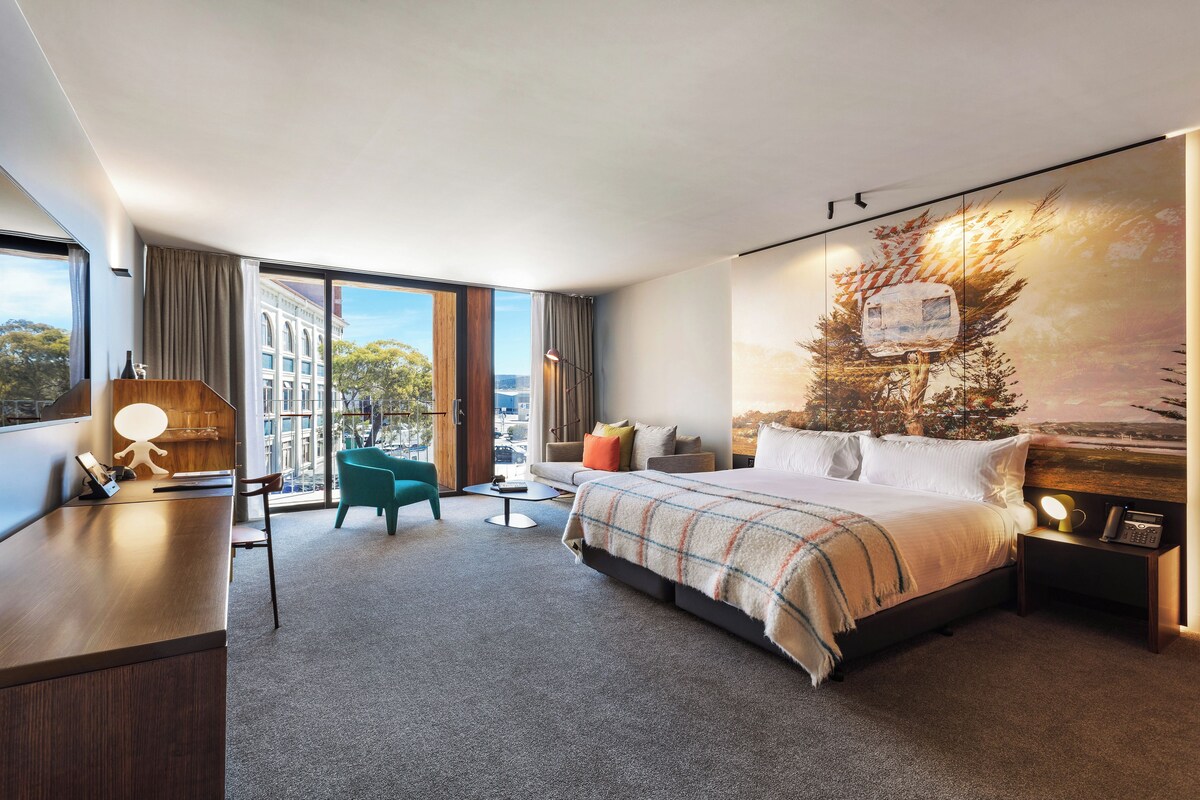 A modern hotel room with a large bed, a desk, a chair, and a view of the city through large windows.