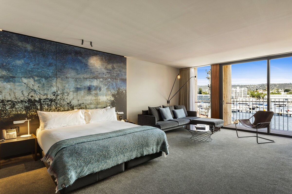 A modern hotel room with a large bed, a sofa, a chair, and a view of the marina.