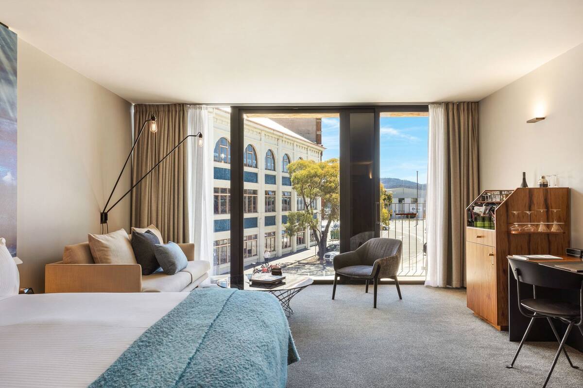 Quarto de hotel com cama, sofá, poltrona, escrivaninha, minibar e vista para a cidade.