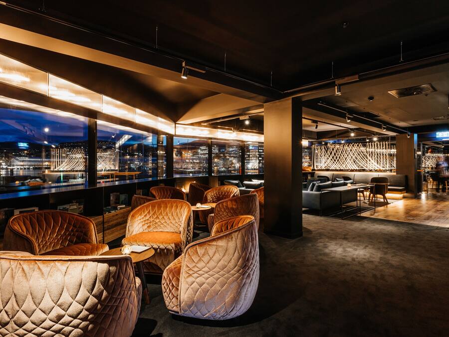 A modern lounge area with cushioned seating, large windows offering city views, and a bar area with glass partitions.