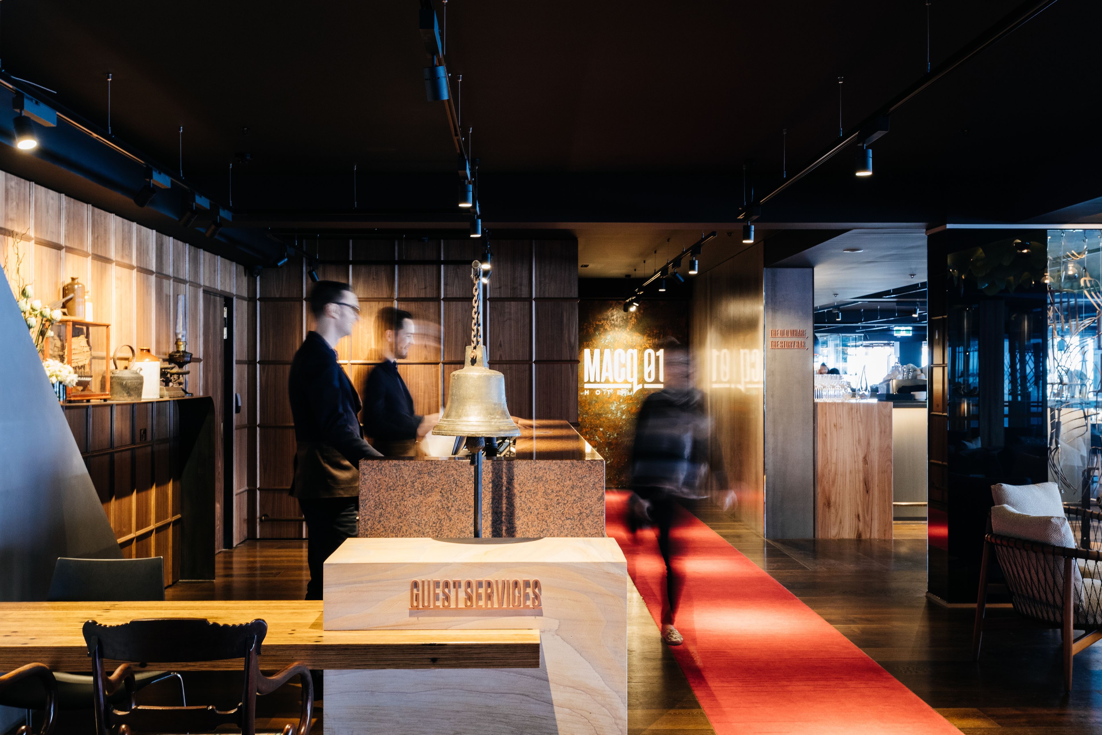 A modern hotel lobby with a wooden reception desk labeled "Guest Services," a red carpet, and a bar area in the background.