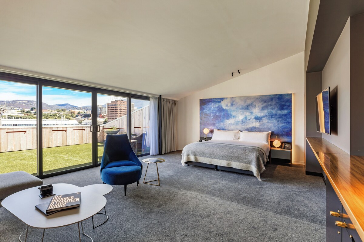 A modern hotel room with a large bed, a seating area with a chair and a small table, and a balcony with a view of the city and mountains.