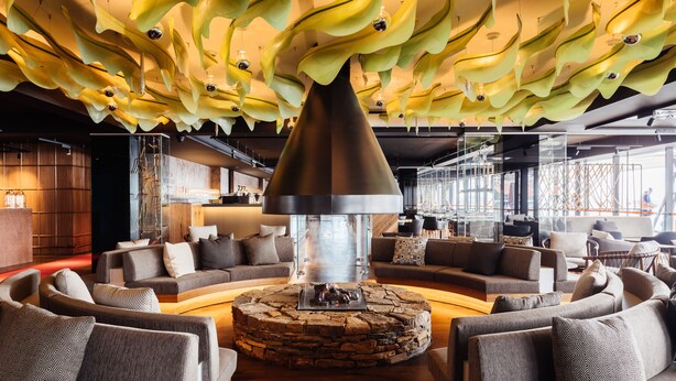 A modern lounge area with sofas arranged around a central stone fireplace, a unique ceiling design with hanging elements, and a view of the outside through large windows. - MACq 01 Hotel (Hobart)