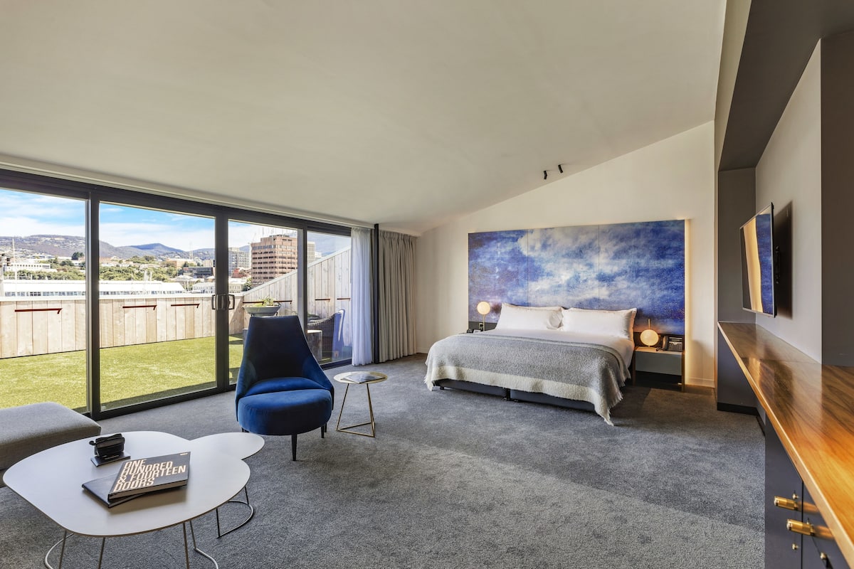 Quarto de hotel moderno com uma grande porta de vidro deslizante que dá para uma varanda, uma cama com vista, uma pequena mesa redonda e uma poltrona azul.