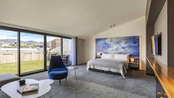 Una habitación de hotel moderna con una cama grande, un área de descanso con una silla y una mesita, y un balcón con vistas a la ciudad y a las montañas.