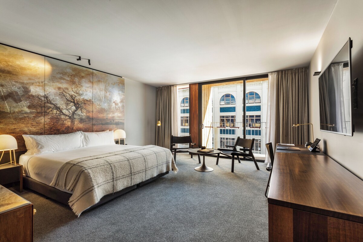 A hotel room with a bed, a desk, a chair, a TV, and a large window with curtains.