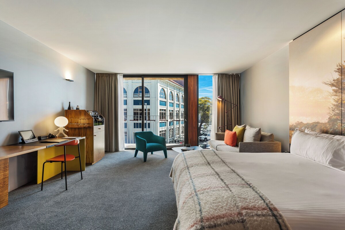 A hotel room with a bed, desk, chair, and a view of buildings through a large window.