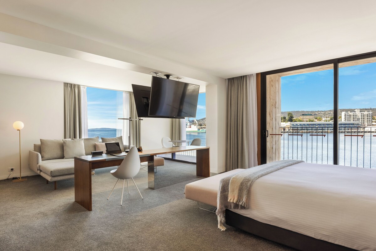 A hotel room with a bed, a desk, a chair, a TV, and a view of the sea.