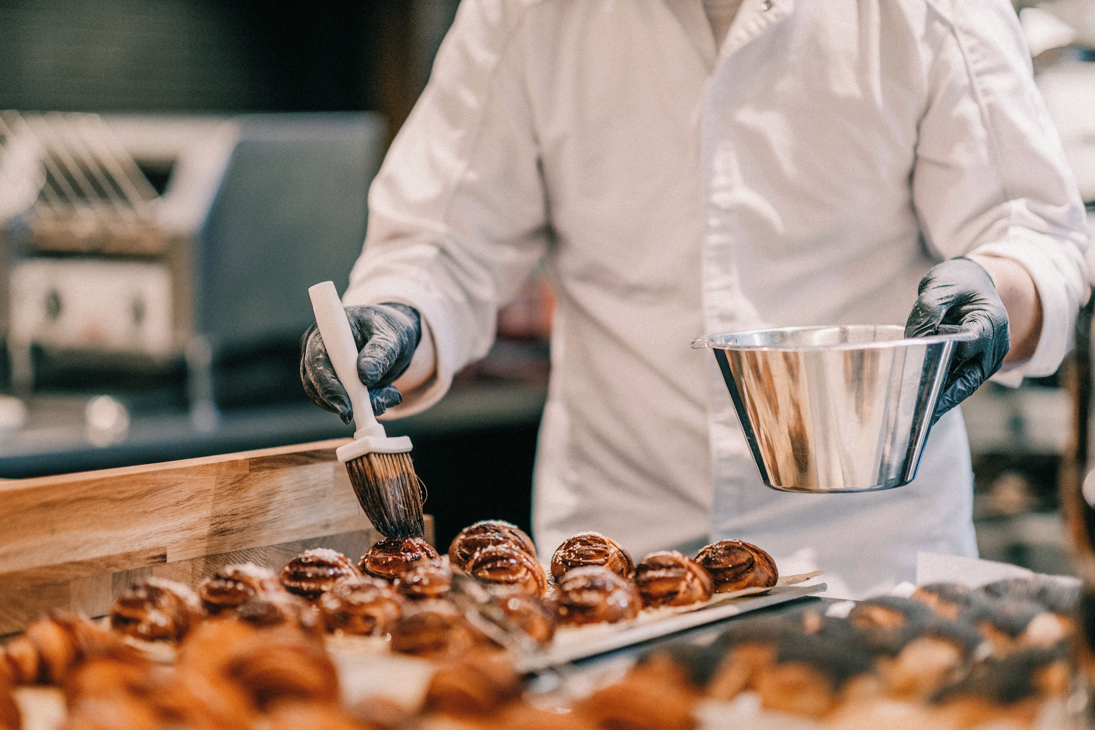 A person in a white coat and gloves, holding a metal container and a brush, painting or applying something to pastries on a tray.