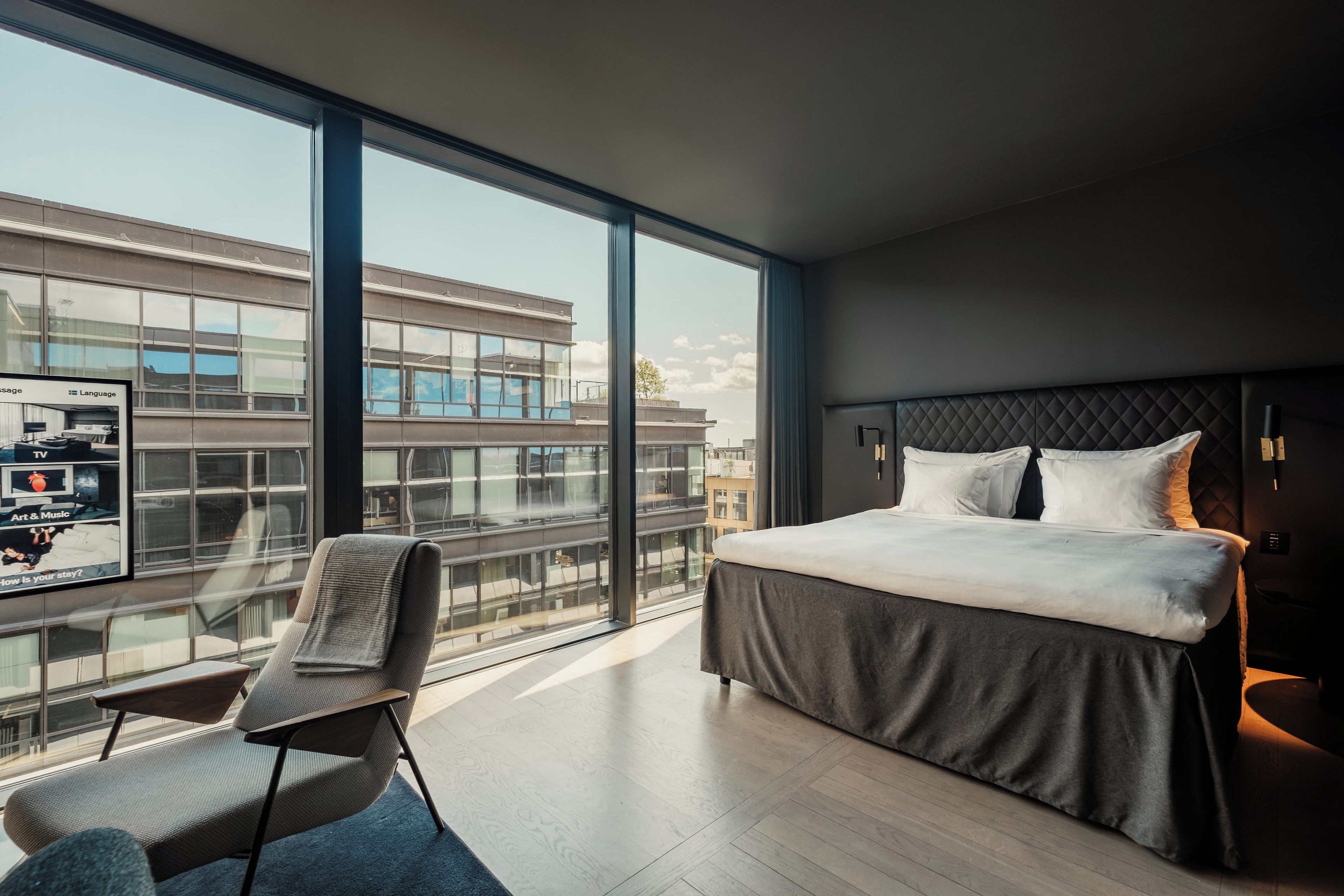A modern hotel room with a large window, a bed, a chair, and a view of the cityscape.