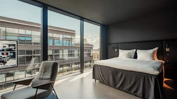 A modern hotel room with a large window, a bed, a chair, and a view of the cityscape.