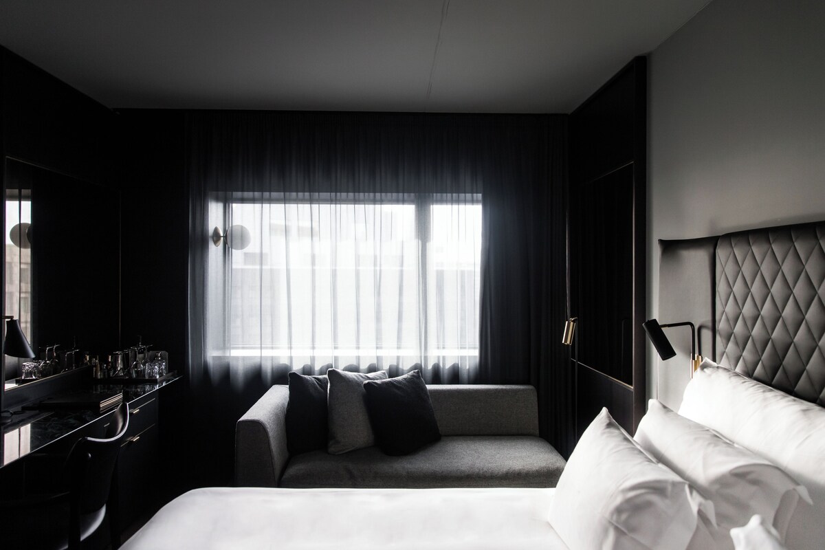 A modern hotel room with a large bed, a desk with a lamp, and a view of the city through sheer curtains.