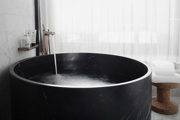 Une grande baignoire noire remplie d’eau, une étagère en bois avec des articles de toilette et une serviette blanche posée sur un tabouret.