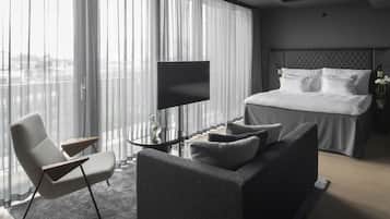 Habitación de hotel moderna con una cama grande, un sofá, un televisor y vistas de la ciudad a través de cortinas translúcidas.