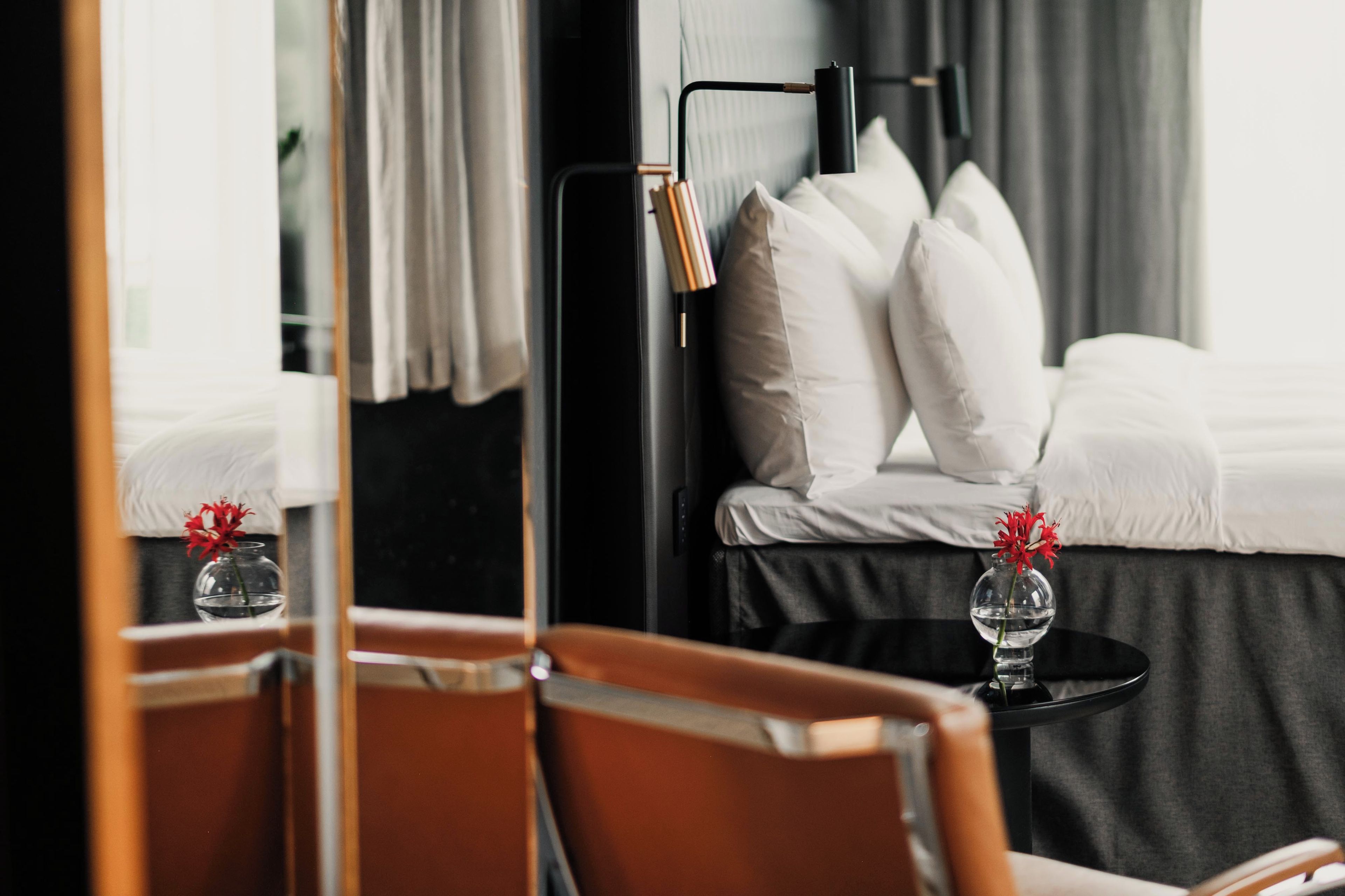 Una camera d'albergo con un letto, un comodino con un vaso di fiori, una sedia e uno specchio che riflette l'ambiente.