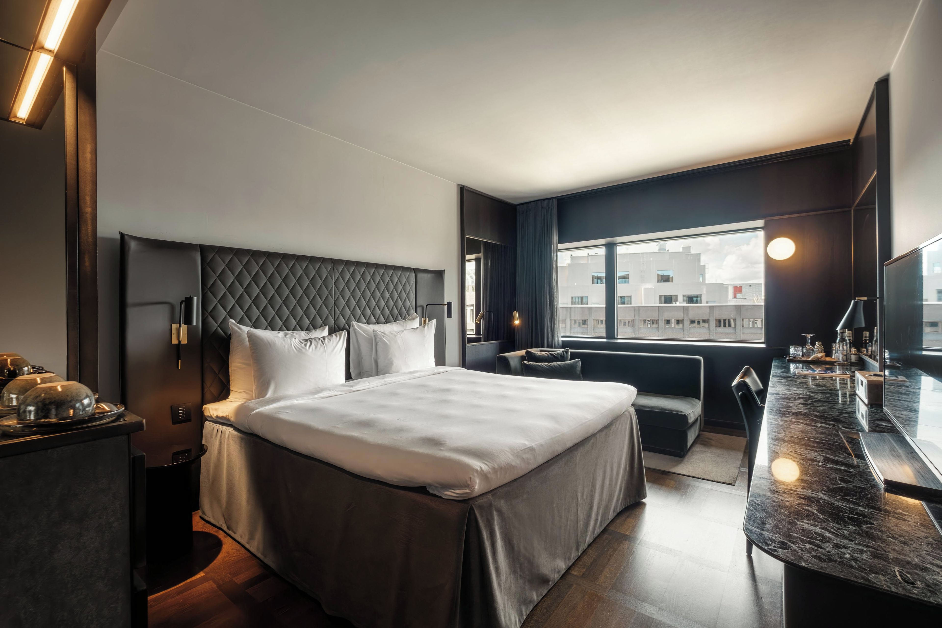 A modern hotel room with a large bed, a desk, and a view of the cityscape through the window.