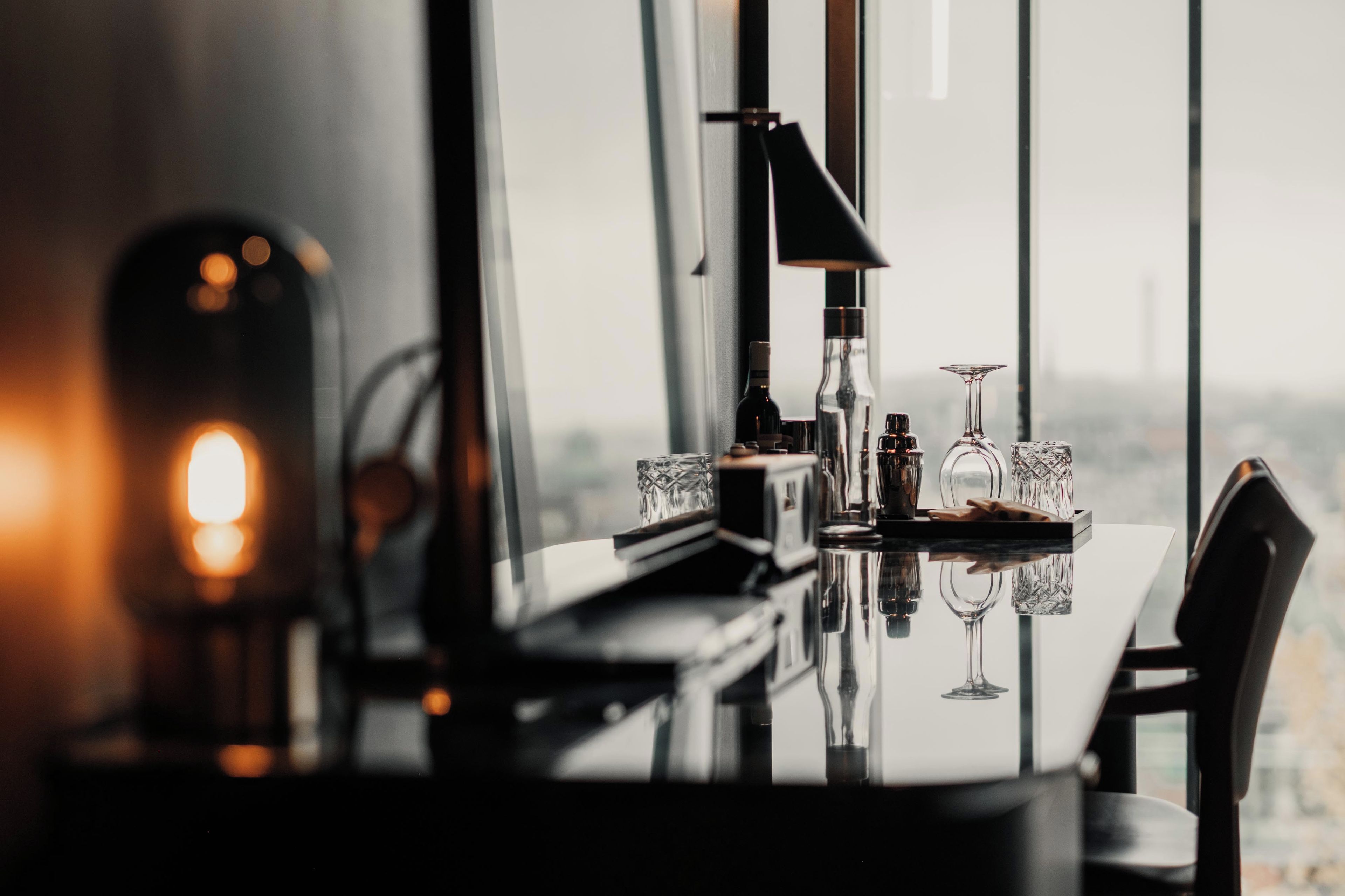 A dining table with a reflective surface, a wine glass, and a bottle, set against a window with a view of a cityscape.