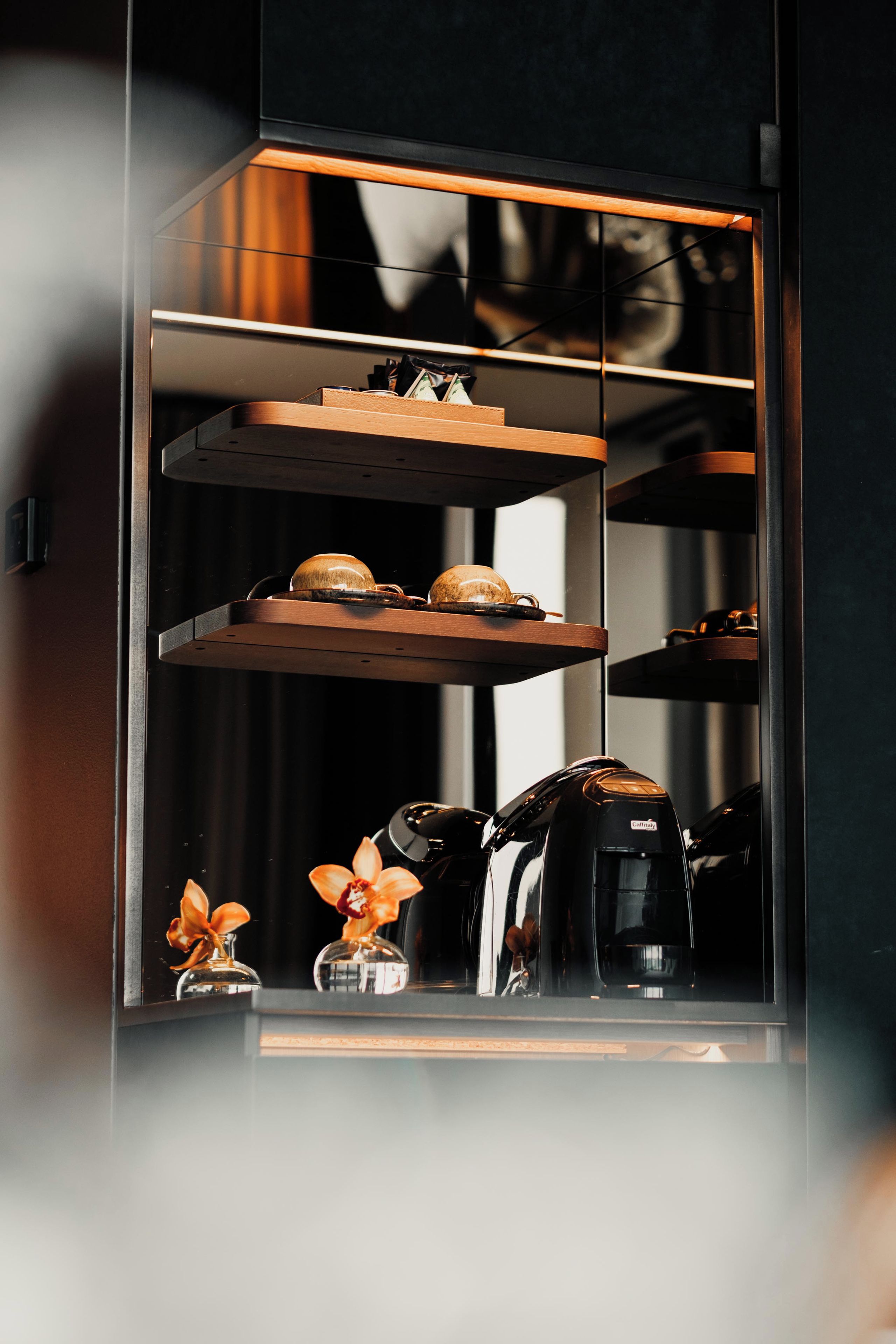 A kitchen with a coffee machine, a shelf with a cup, and a vase with flowers.