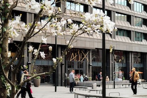 A modern building with a glass facade, outdoor seating, and a large planter with flowers. - At Six (Stockholm)