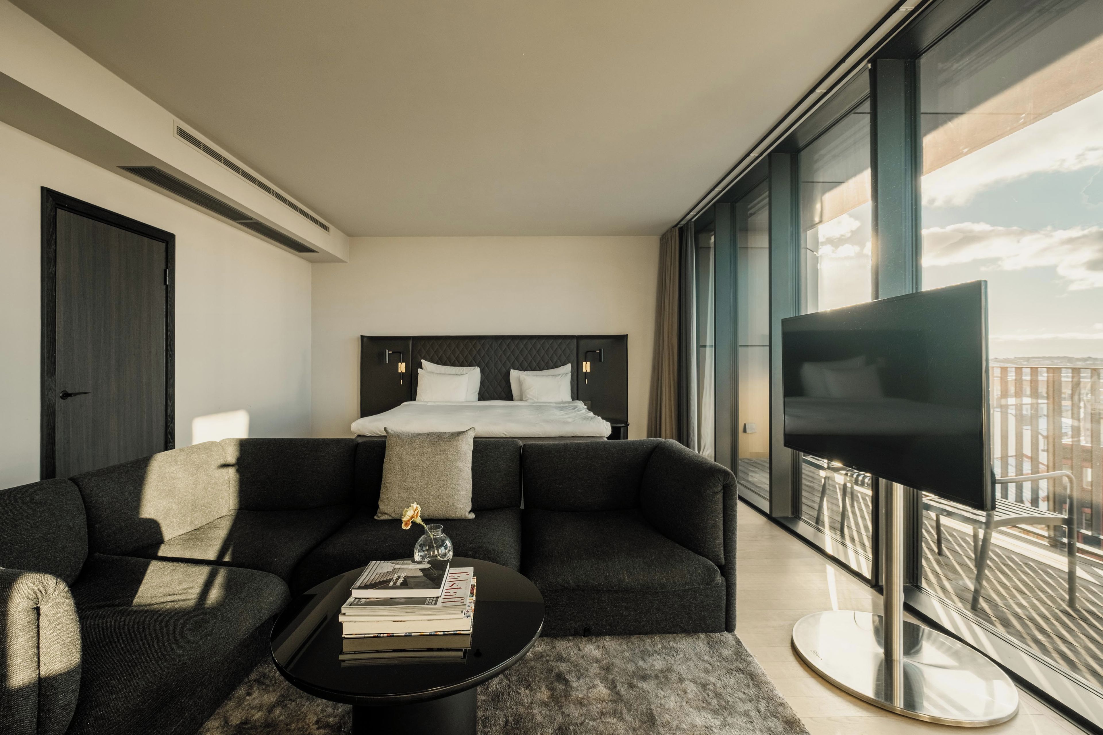 A modern hotel room with a large bed, a sofa, a coffee table with books, and a TV.