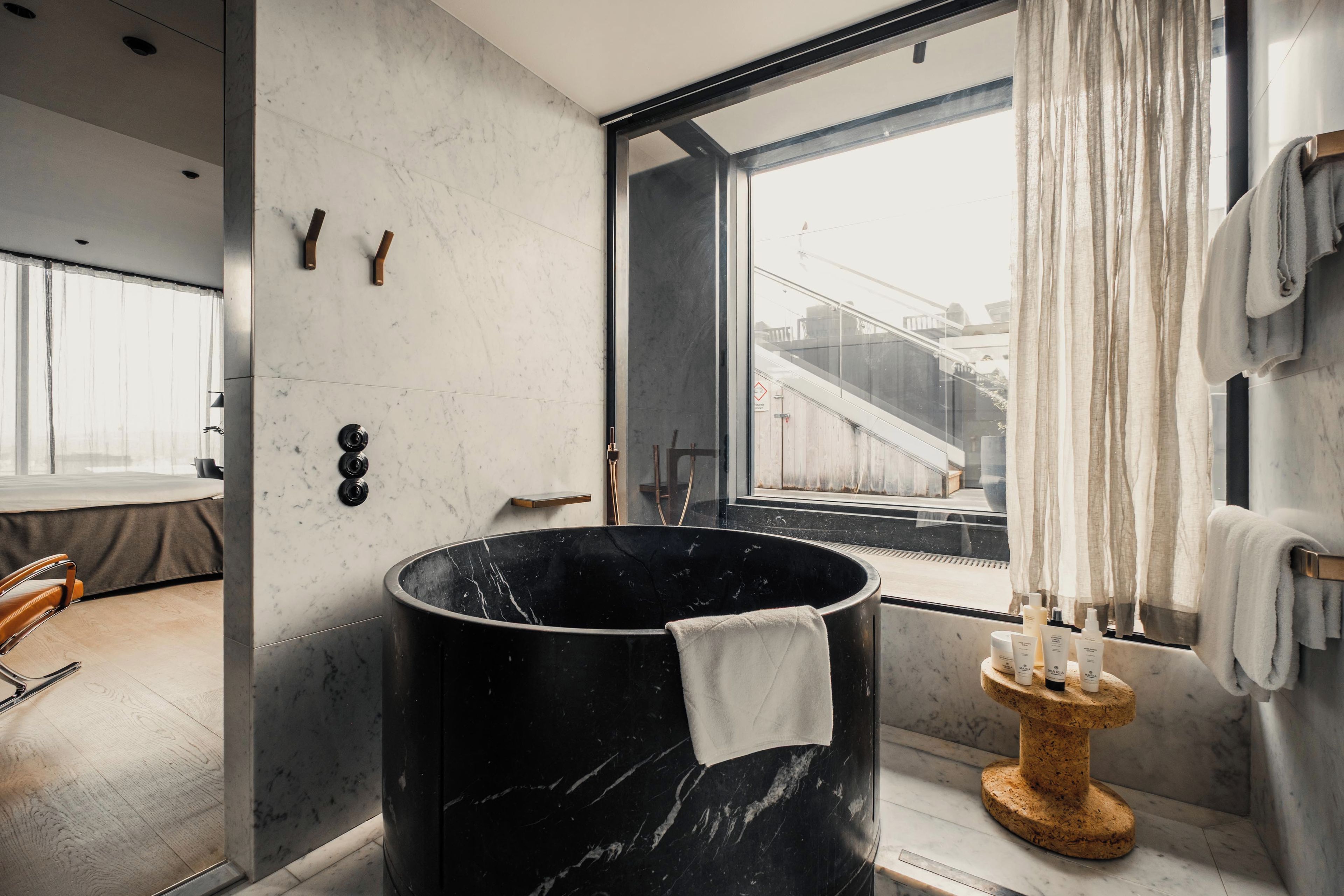 A modern bathroom with a large black marble tub, a wooden towel rack, and a view of the city through a large window.