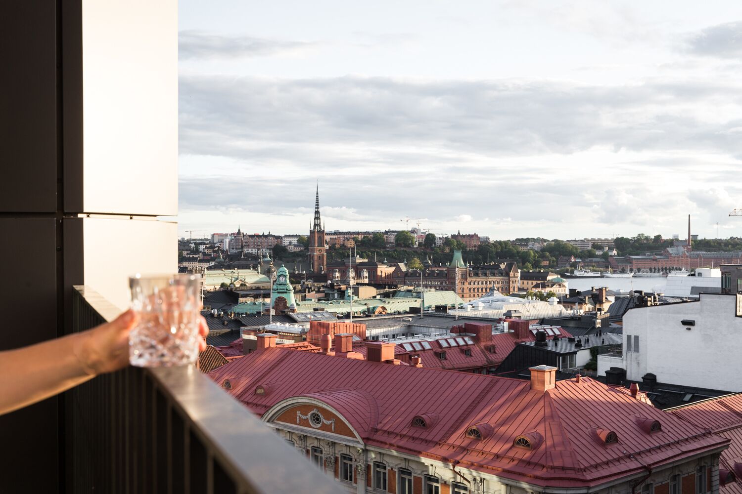 Une vue urbaine avec un clocher d’église imposant, des bâtiments aux toits rouges et un balcon sur lequel se trouve un verre d’eau.