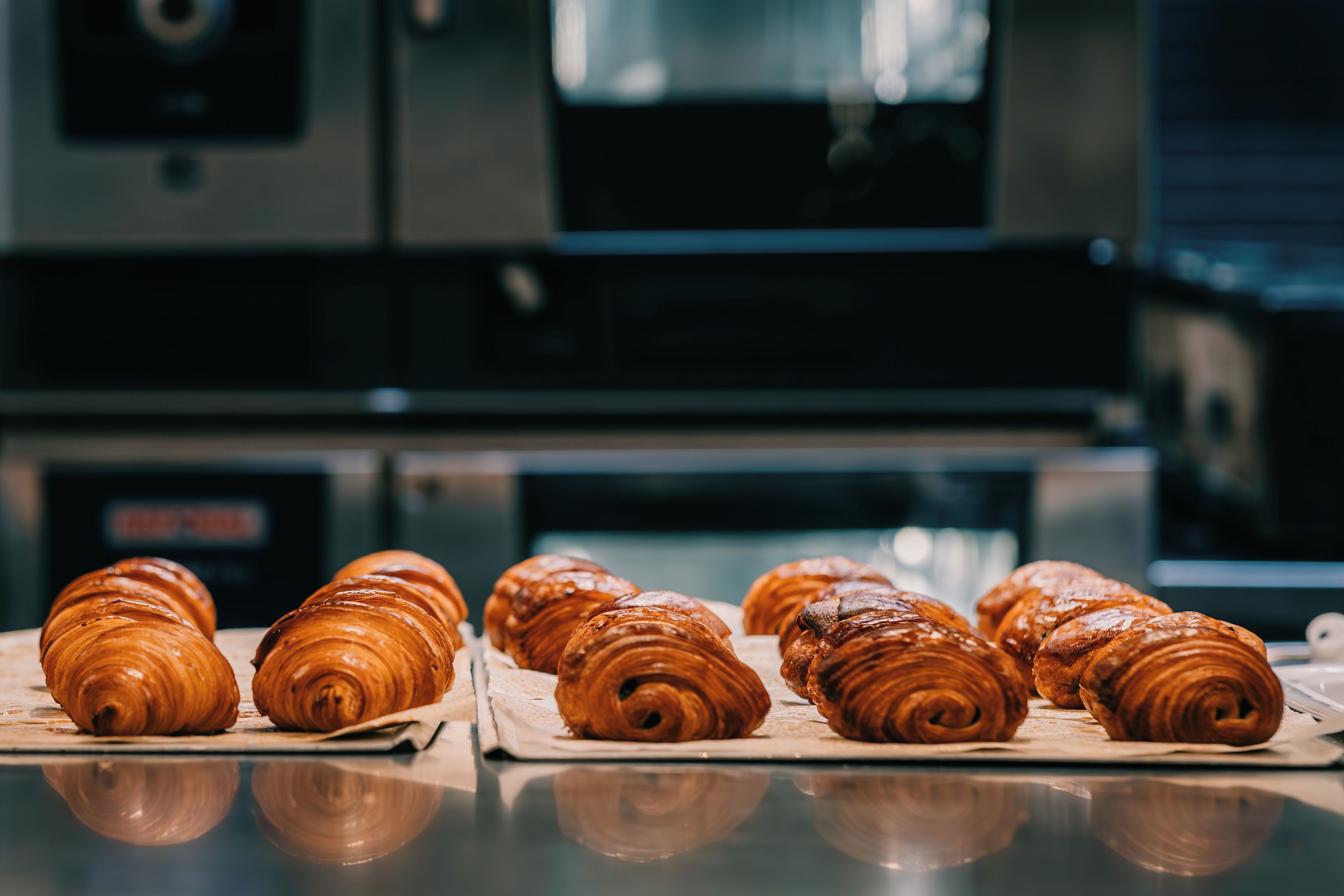 Eine Reihe Croissants auf einer spiegelnden Fläche, im Hintergrund ein Küchengerät.