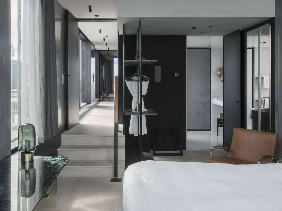A modern hotel room with a large bed, a seating area, a vanity, and a view of the hallway.