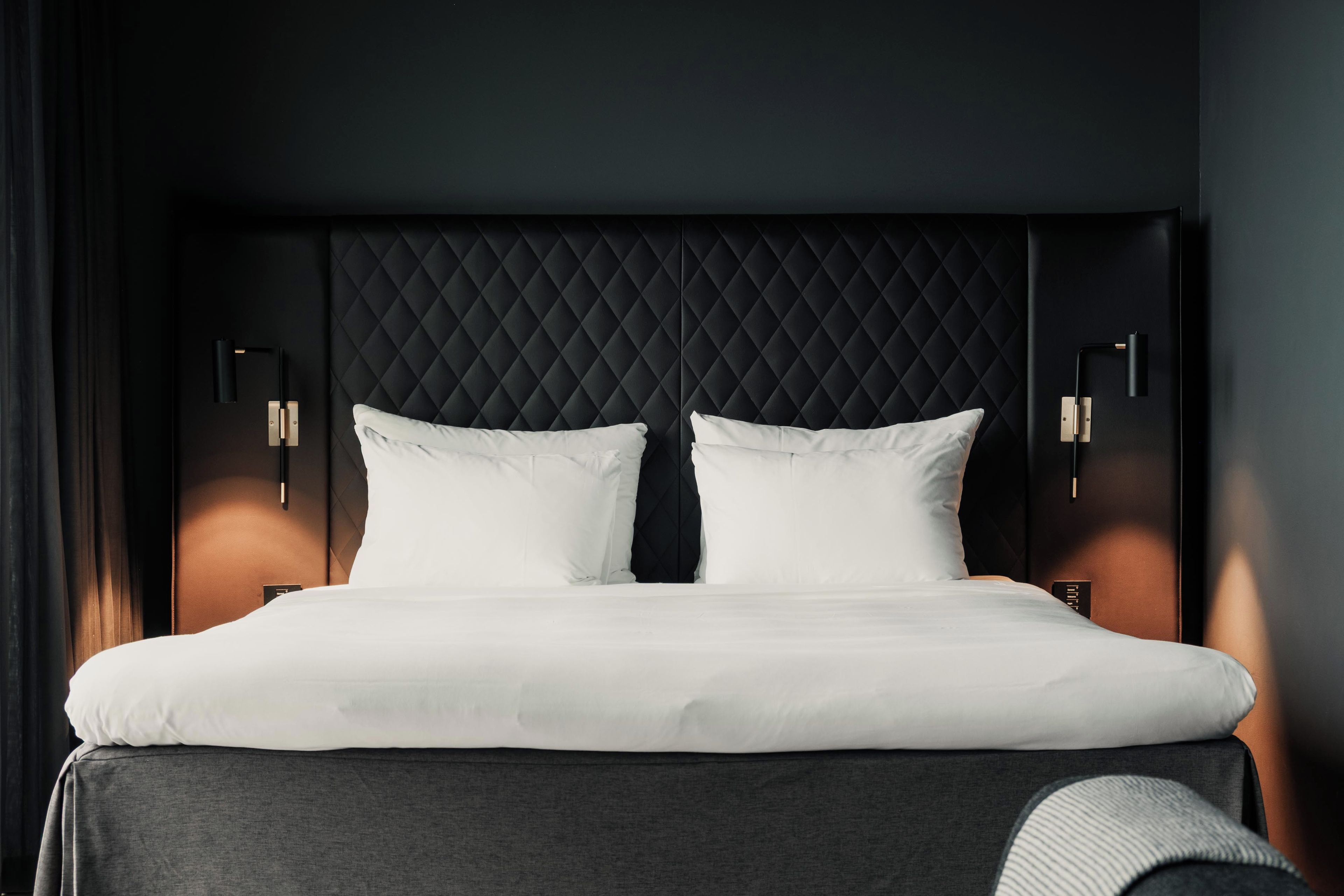 A modern hotel room with a large bed, quilted headboard, and wall-mounted bedside tables.